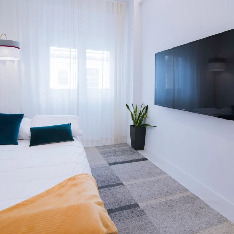 Modern bedroom with large TV, white walls, bed with blue pillows, plant